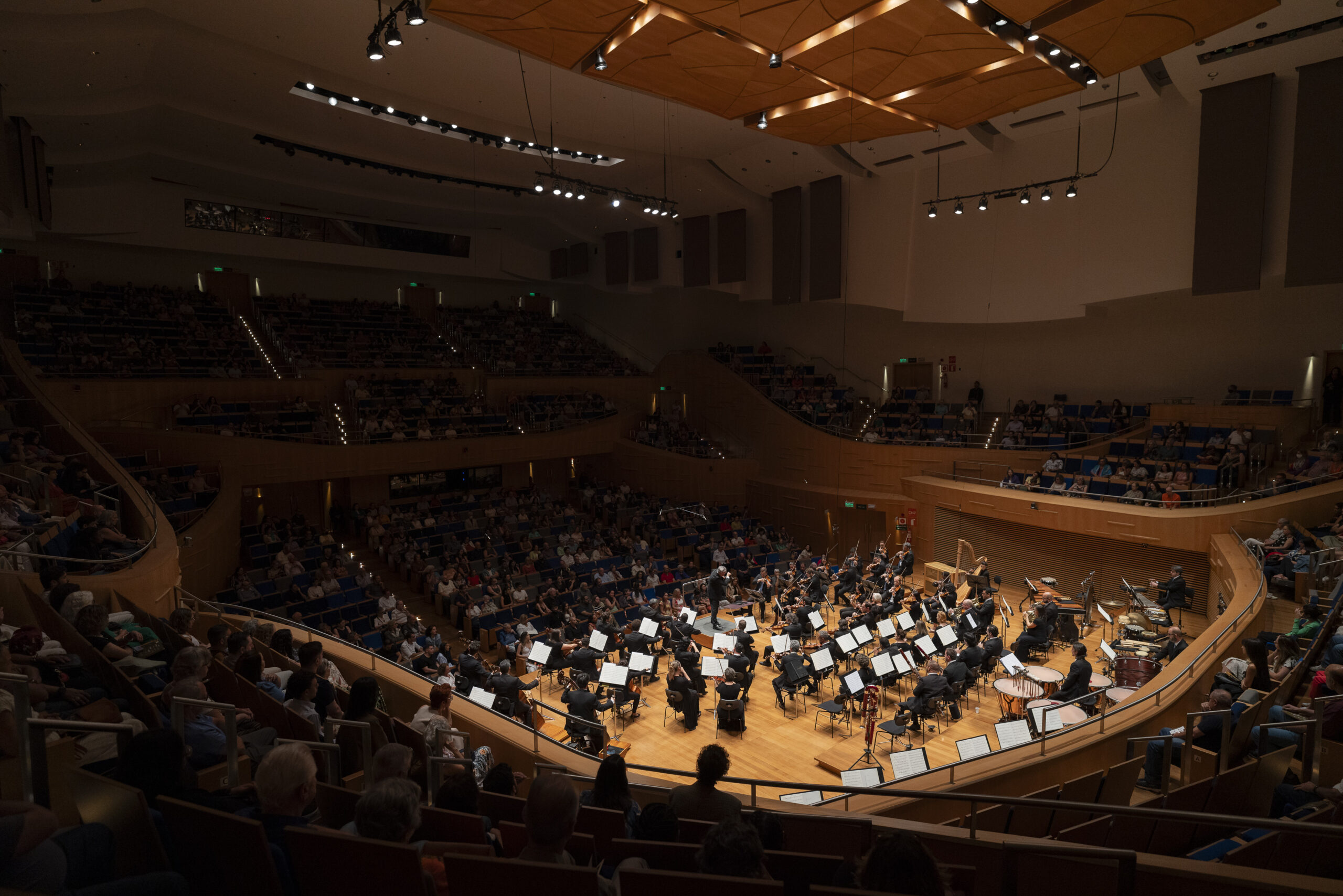 Foto do interior da sala de concertos da Sala Minas Gerais. Ela mostra a visão de quem está no balcão palco e vê o maestro e as outras plateias de frente, mas os músicos de costas.