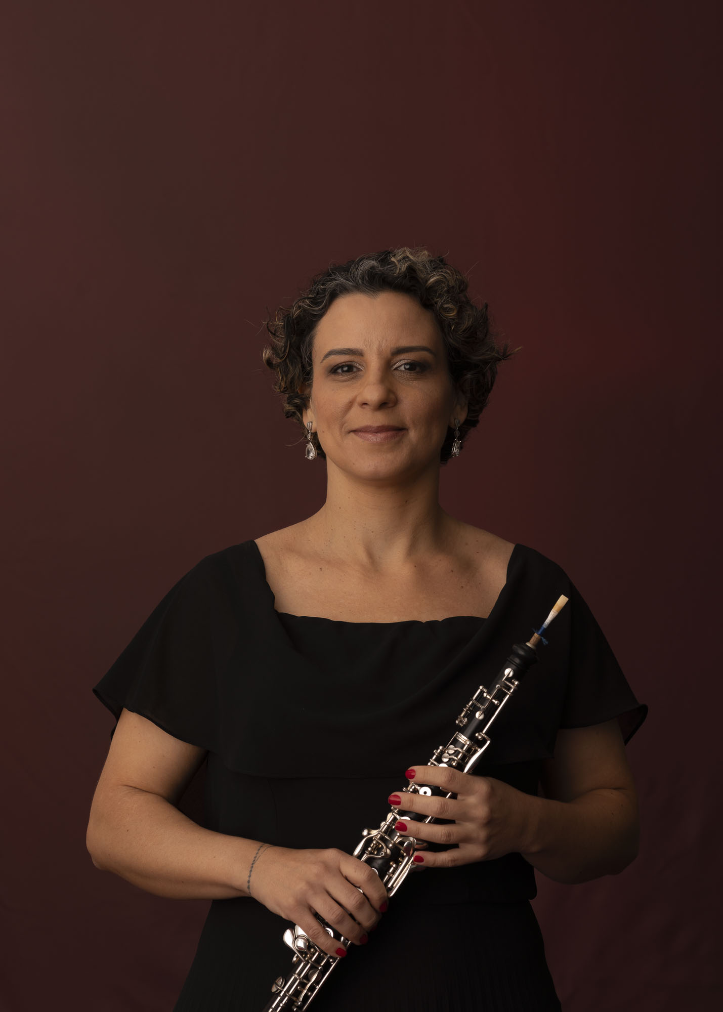 Foto da oboísta Maria Fernanda. Ela está olhando para frente com o oboé nas mãos e o fundo é um tecido vinho.