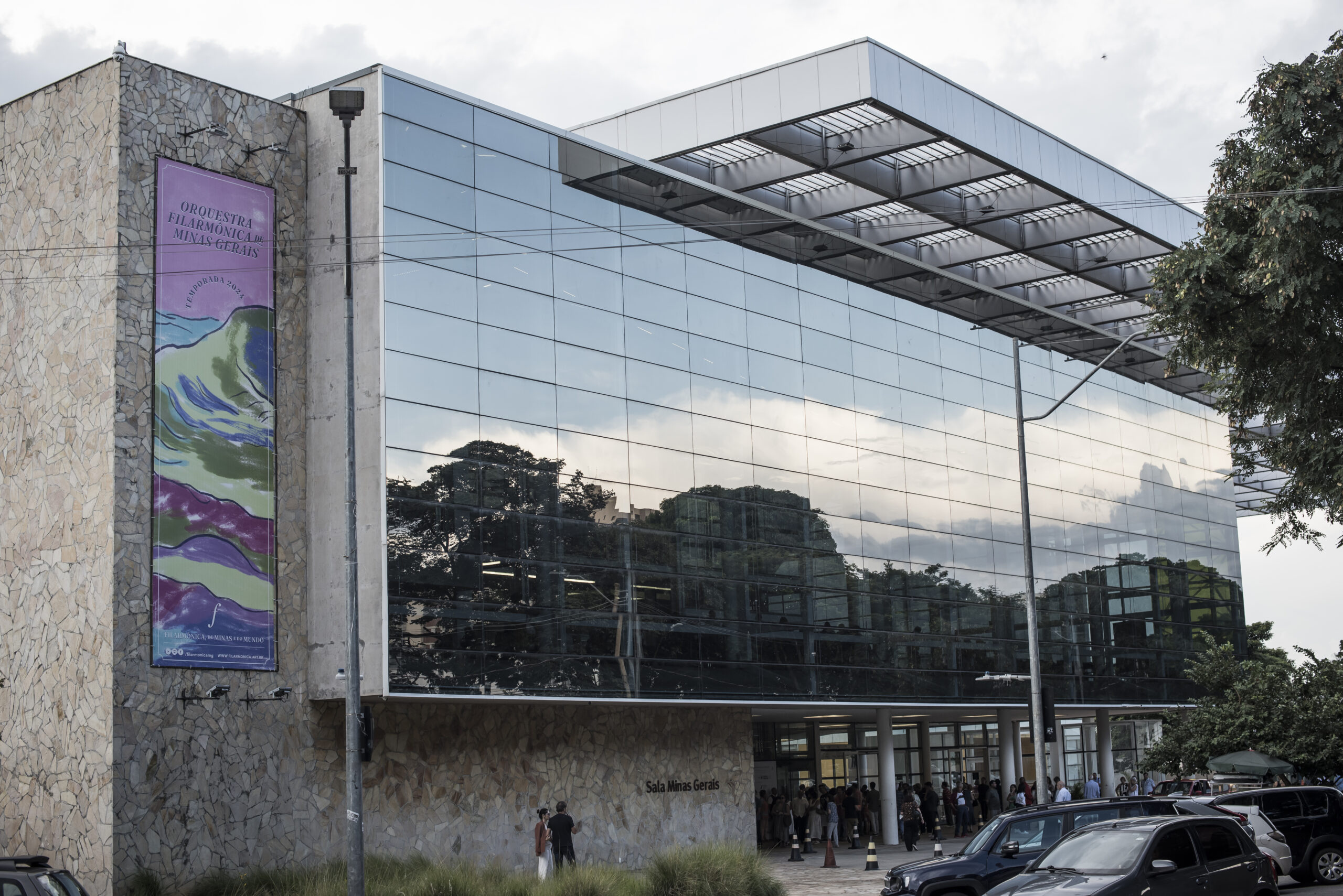Foto diurna da fachada do lado de fora da Sala Minas Gerais. Você pode perceber a frente espelhada que reflete as nuvens do céu