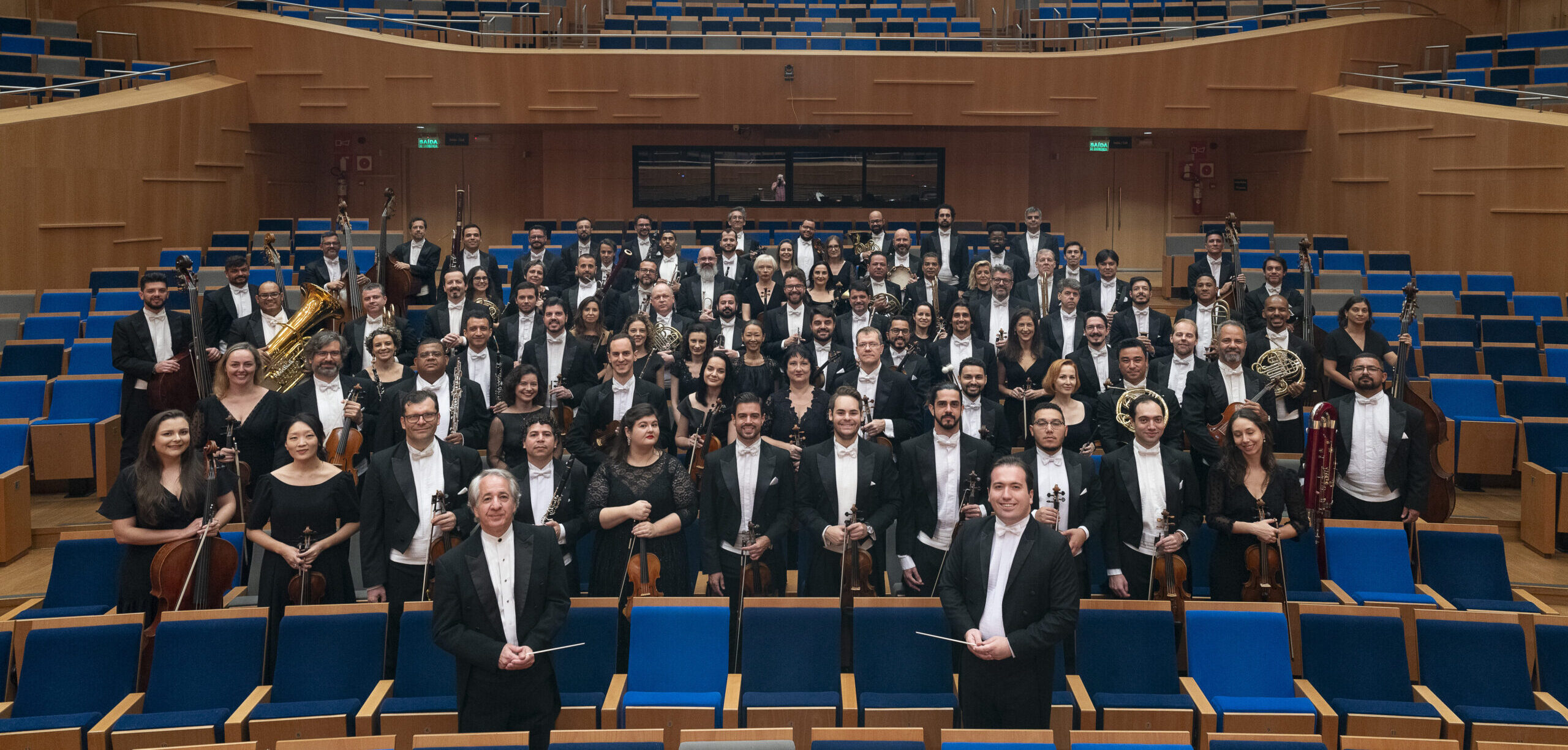 Foto dos músicos da Filarmônica e de seus dois maestro. Eles estão de pé na plateia, olhando para a câmera e segurando seus instrumentos. Foto de Eugênio Sávio. 2025