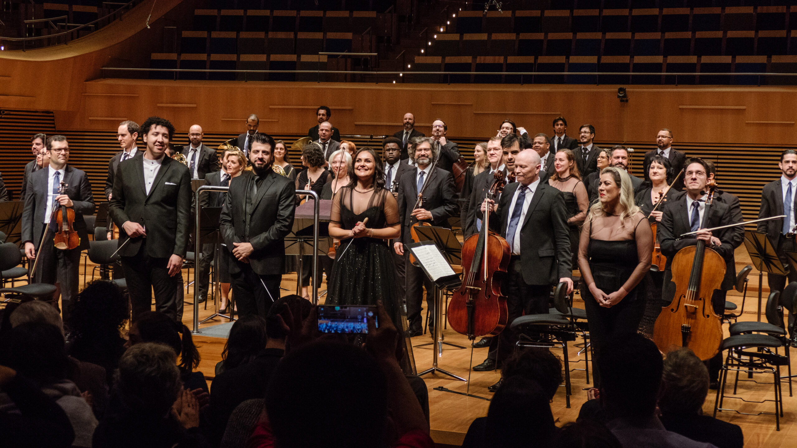 Foto mostrando os quatro regentes ativos do laboratório de regência em cima do palco. Eles estão recebendo os aplausos do público ao final do concerto. Foto de Felipe GIubilei.