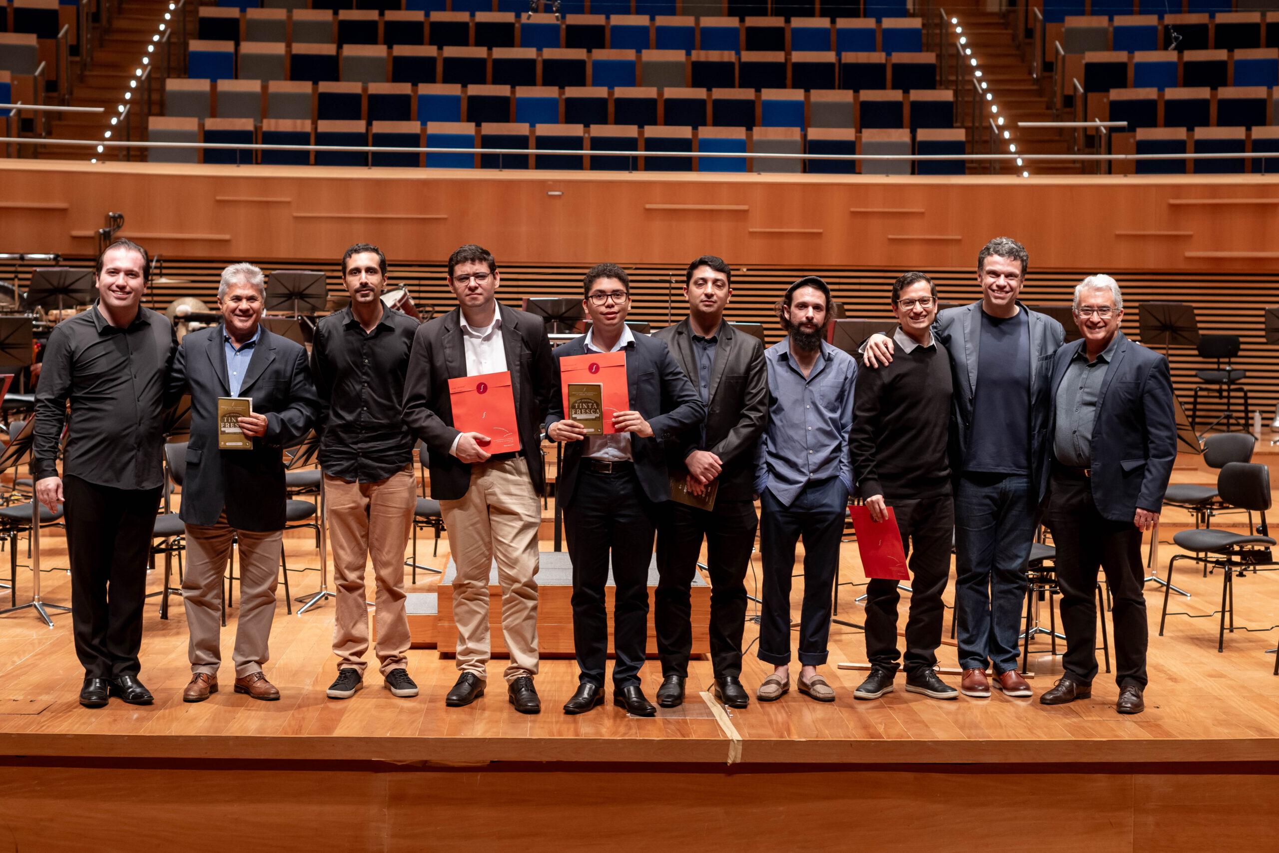 Foto dos compositores que participaram do Festival Tinta Fresca. Eles estão em cima do palco com seus certificados na mão. Entre eles estão os jurados e o palco está vazio já sem músicos. Foto de Alexandre Rezende